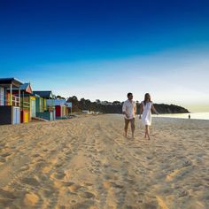 a man and woman walking on the beach in front of some colorful huts with water behind them