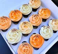 twelve cupcakes with orange and white frosting in a box on a table