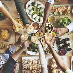 many people are holding wine glasses over a table full of food