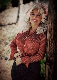 a woman with blonde hair wearing a red shirt and black pants, leaning against a wooden pole