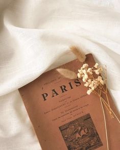 an old book is laying on a bed with white sheets and flowers in front of it