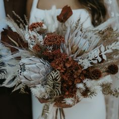 a bride holding a bouquet of dried flowers and feathers in her hands with the groom standing behind her