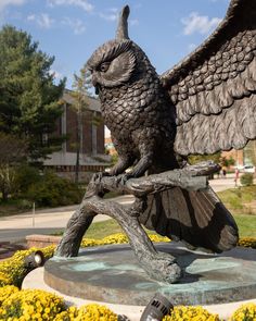 a statue of an owl perched on a branch with its wings spread out, in front of yellow flowers