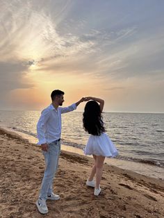 a man and woman standing on top of a sandy beach next to the ocean at sunset