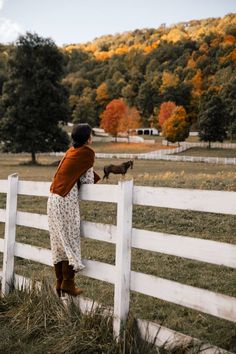 a woman leaning on a white fence and petting a small dog in the field