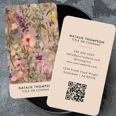 two business cards sitting on top of a black plate next to a gray background with pink, yellow and purple flowers