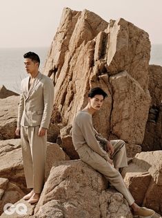 two men in suits sitting on rocks near the ocean and one man standing next to them