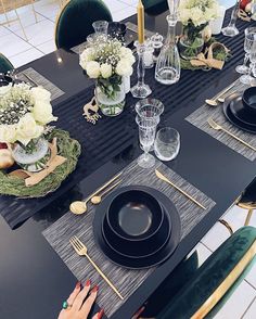 a table set with black plates and silverware, gold candlesticks and white flowers