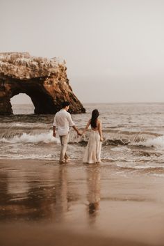 a man and woman holding hands walking on the beach with waves crashing in front of them