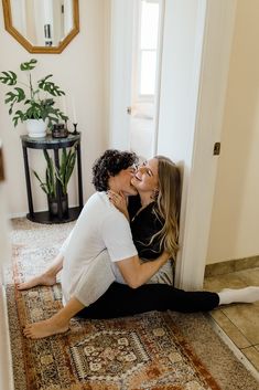 two women sitting on the floor kissing each other in front of a door with a potted plant behind them