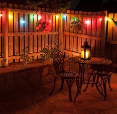 an outdoor table and chairs are lit up with christmas lights on the fence behind them