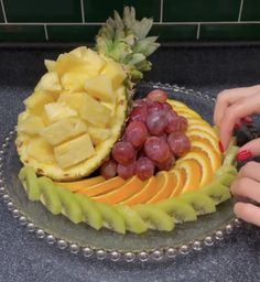 a person is cutting up fruit on a glass platter with grapes, pineapples and oranges