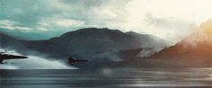 an airplane is flying over the water with mountains in the background and clouds above it