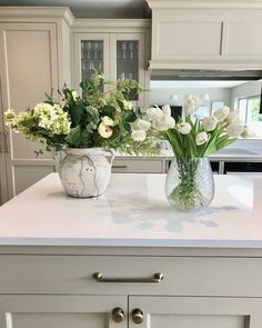 An off-white kitchen island with white countertops is styled with farmhouse glass vases. One is a white brushed stone urn with hydrangeas, eucalyptus, and ferns. The other is a clear crystal glass vase with white tulips. Glass cabinetry in the backdrop reveals an assortment of coordinating crystal glass drinkware...   Image: roombyroomdublin