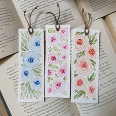 two bookmarks with watercolor flowers on them are hanging from twine strings in front of an open book