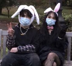 two people sitting on a bench wearing bunny ears, masks and black clothing with one person holding up the peace sign