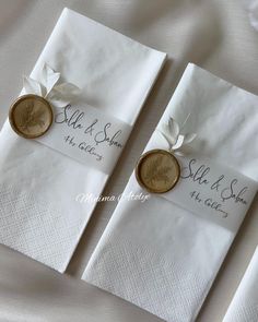 two white napkins with gold buttons and ribbons on them are sitting next to each other