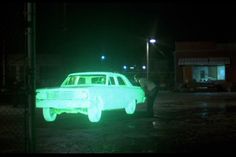 a green car is parked in the rain at night with people walking by and one person standing near it