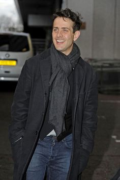 a man in a black coat and scarf smiles at the camera as he stands outside