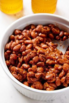 a white bowl filled with baked beans next to two glasses on a counter top and one has a spoon in it