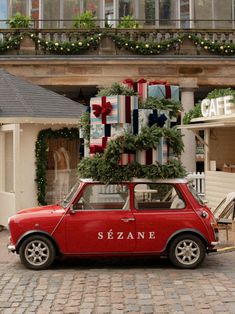 a small red car parked in front of a building with christmas presents on it's roof