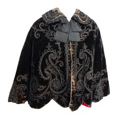 Edwardian black velvet cape with soutache trim on collar and throughout body. Silk neck bow. Hook and eye closure along front. Re-lined in red cotton Luxury Black Medieval Outerwear, Luxury Medieval Style Black Outerwear, Luxury Medieval Black Outerwear, Luxury Black Gothic Cape, Elegant Velvet Outerwear For Costume, Black Velvet Costume Outerwear, Velvet Cape, Black Cape, Capes For Women