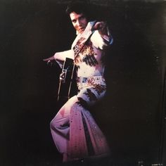 a man with a guitar in his hand and an outfit on, standing up against a black background