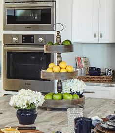 three tiered trays hold apples and lemons in front of an oven with white flowers on the counter