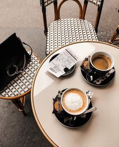 two cups of coffee sitting on top of a white table next to a black purse