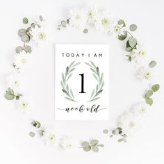 a card with the number one on it surrounded by white flowers and greenery, which reads today i am