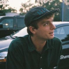 a young man wearing a hat and looking off to the side in front of parked cars