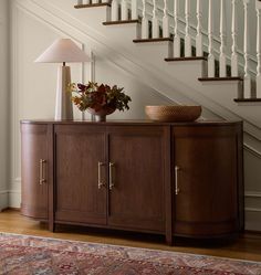 a wooden cabinet sitting under a stair case next to a table with a lamp on it