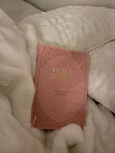 a pink bible laying on top of a white bed covered in fluffy blankets and pillows