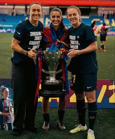 two women standing next to each other with a trophy in front of them on a soccer field