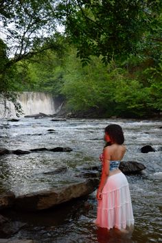 a woman standing in the water next to a waterfall wearing a pink and white dress