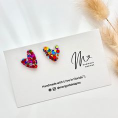 two colorful heart shaped earrings sitting on top of a white card next to some dry grass