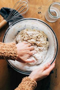 two hands mixing dough in a bowl on top of a wooden table next to utensils