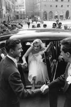 two men shaking hands in front of a car with a woman standing next to them