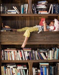 a young boy laying on top of a wooden book shelf filled with lots of books