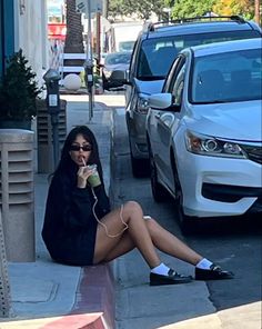 a woman is sitting on the sidewalk talking on her cell phone and drinking from a cup