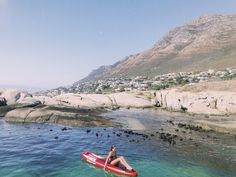 a person on a red kayak in the water near some rocks and houses with mountains in the background