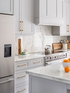 a white kitchen with marble counter tops and stainless steel appliances in the center, along with an orange bowl on the island