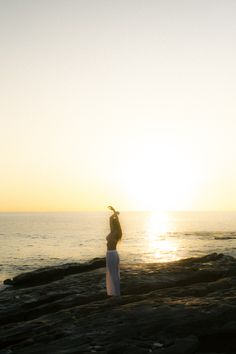 a woman standing on top of a rock covered beach next to the ocean at sunset