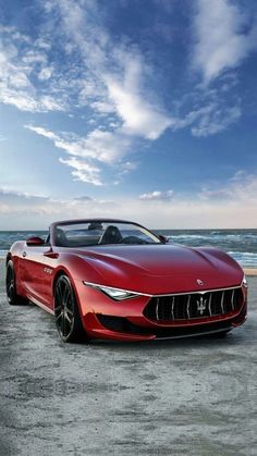 a red masera parked on the beach next to the ocean with clouds in the sky