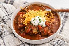 a white bowl filled with chili and meat topped with sour cream on top of a checkered towel