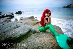 a woman with long red hair sitting on rocks near the ocean wearing a green mermaid tail