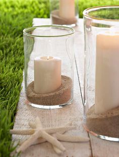 three candles are sitting in glass vases on the ground with sand and starfish