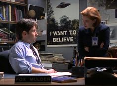 two people standing in an office with books on the desk and posters on the wall behind them