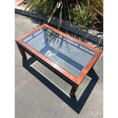 a wooden table with glass top sitting in the middle of a parking lot next to plants