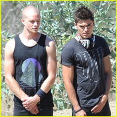 two young men standing next to each other in front of trees and bushes, one wearing a black shirt